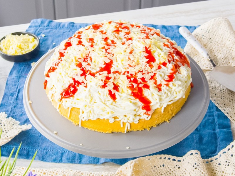 Un pastel helado de espagueti en bandeja está sobre una mesa de madera. A su izquierda hay algunas fresas. A su derecha, una rebanadora de pastel.
