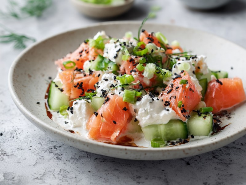 Un plato con ensalada de pepino con queso cottage y salmón. Todo ello adornado con sésamo negro, cebolletas y eneldo.